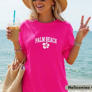May include: A bright pink t-shirt with the text "PALM BEACH" in white, along with a white flower graphic. The shirt is worn with a straw hat, sunglasses, and bracelets. The background shows a beach and ocean.