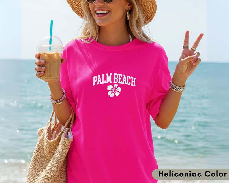 May include: A bright pink t-shirt with the text "PALM BEACH" in white, along with a white flower graphic. The shirt is worn with a straw hat, sunglasses, and bracelets. The background shows a beach and ocean.