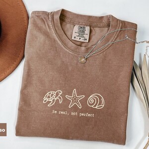 Pode incluir: Uma t-shirt cor de espresso com um gráfico de linhas brancas de uma tartaruga marinha, uma estrela-do-mar e uma concha. Abaixo do gráfico, o texto diz "Be real, not perfect". A t-shirt tem gola redonda e uma etiqueta da marca Comfort Colors.