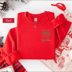 May include: A red crewneck sweatshirt with the text "Tired Nurses Club Sophia" in green and gold lettering. A silver necklace with a sun pendant is visible. The word "Red" is in a red rectangle.