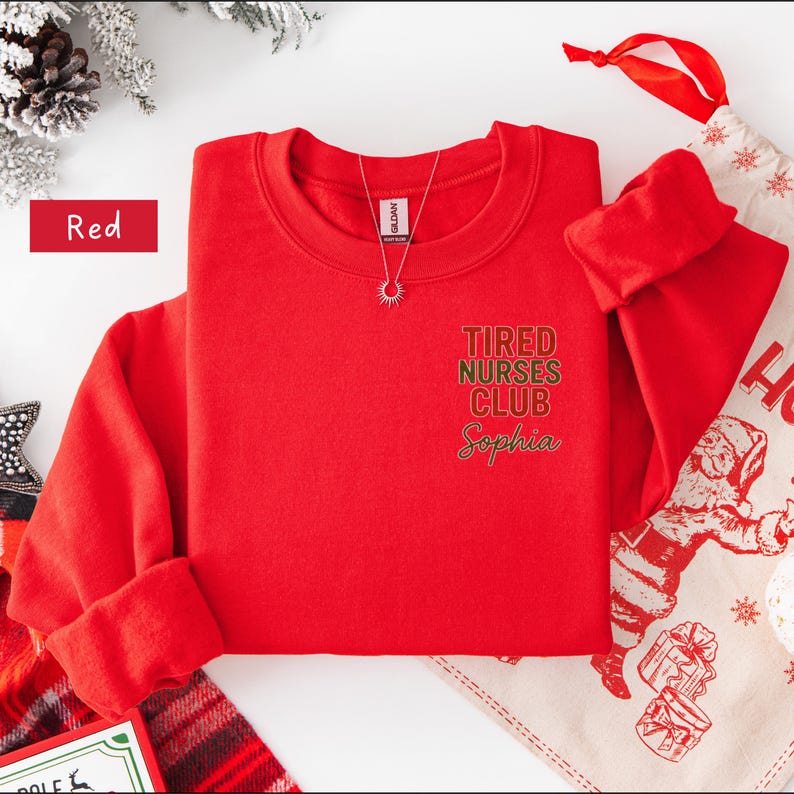 May include: A red crewneck sweatshirt with the text "Tired Nurses Club Sophia" in green and gold lettering. A silver necklace with a sun pendant is visible. The word "Red" is in a red rectangle.