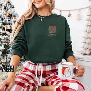 May include: Forest green sweatshirt with the text "Tired Nurses Club Sophia" in red and green. The person is wearing red and white plaid pajama pants and holding a mug with a Santa Claus design.