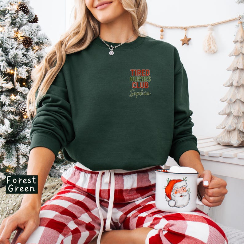 May include: Forest green sweatshirt with the text "Tired Nurses Club Sophia" in red and green. The person is wearing red and white plaid pajama pants and holding a mug with a Santa Claus design.