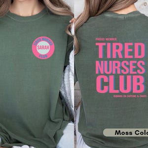 May include: Moss green t-shirt with the text "TIRED NURSES CLUB" in pink and blue on the back. The front features a circular design with "TIRED NURSES CLUB" and the name "SARAH". The shirt is a comfortable fit.