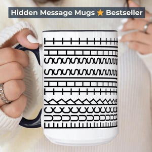 Puede incluir: Taza de cerámica blanca con asa y borde negros, con un patrón geométrico negro repetido. La taza tiene el texto "Hidden Message Mugs Bestseller" en la parte superior. La taza está sujeta por una persona.