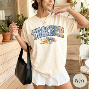 以下が含まれることがあります： アイボリーの半袖Tシャツ。青とオレンジの花柄とギンガムチェックのパターンで「TEACHER」という文字がプリントされています。文字の下には青いリボンが付いた黄色い鉛筆のグラフィックがあり、鉛筆には筆記体で「Mrs. Emily」と書かれています。