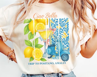 May include: Ivory-colored t-shirt with a graphic featuring lemons, a cocktail, and a blue scooter. The text "Ciao Bella" and "Trip to Positano, Amalfi" are displayed. The design includes a blue and yellow floral pattern.