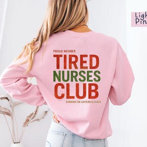 May include: Light pink sweatshirt with the text "TIRED NURSES CLUB" in red and green. The phrase "RUNNING ON CAFFEINE & CHAOS" is below the club name. The words "PROUD MEMBER" are above the club name.