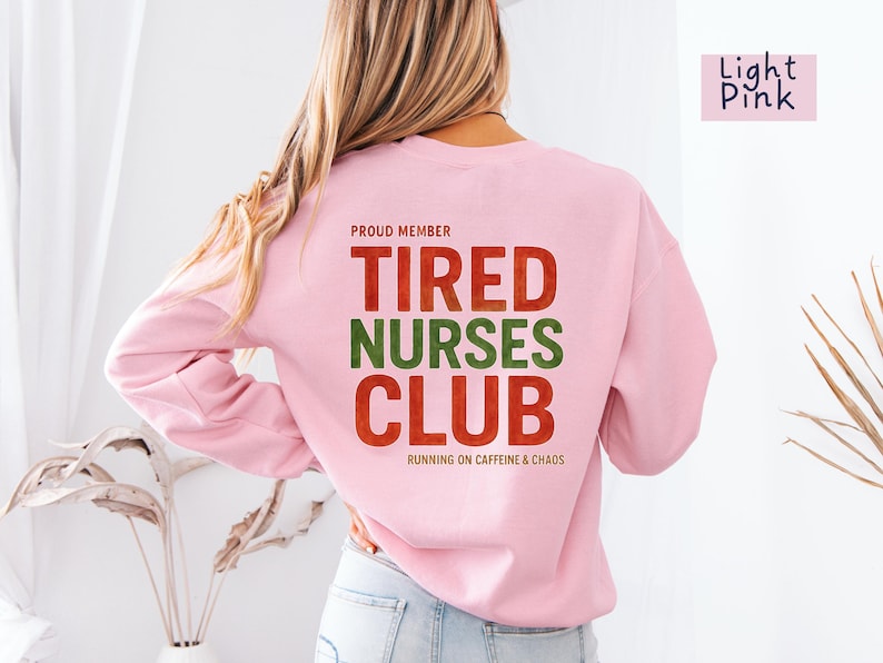 May include: Light pink sweatshirt with the text "TIRED NURSES CLUB" in red and green. The phrase "RUNNING ON CAFFEINE & CHAOS" is below the club name. The words "PROUD MEMBER" are above the club name.
