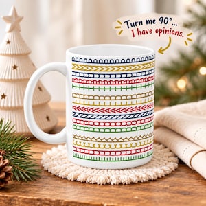 Può includere: Una tazza in ceramica bianca con file orizzontali di motivi geometrici colorati in rosso, blu, verde e oro. Un fumetto sopra la tazza contiene il testo: Turn me 90°... I have opinions. La tazza ha un manico sul lato sinistro.