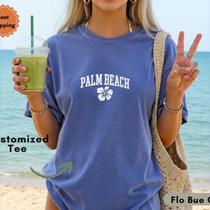 May include: A blue t-shirt with the text "PALM BEACH" in white, along with a white flower graphic. The shirt is a solid color and appears to be made of a soft material. The image also includes a green drink in a clear plastic cup.