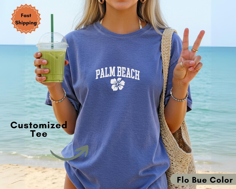 May include: A blue t-shirt with the text "PALM BEACH" in white, along with a white flower graphic. The shirt is a solid color and appears to be made of a soft material. The image also includes a green drink in a clear plastic cup.