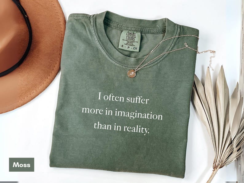 May include: A moss green t-shirt with the text "I often suffer more in imagination than in reality." A gold necklace with a pendant rests on the shirt. A brown hat and dried plant are also in the image.