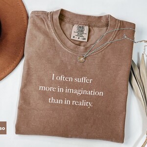 May include: Espresso-colored t-shirt with the quote "I often suffer more in imagination than in reality" in white text. A silver necklace with a pendant and a brown hat are next to the shirt. The shirt has a Comfort Colors tag.