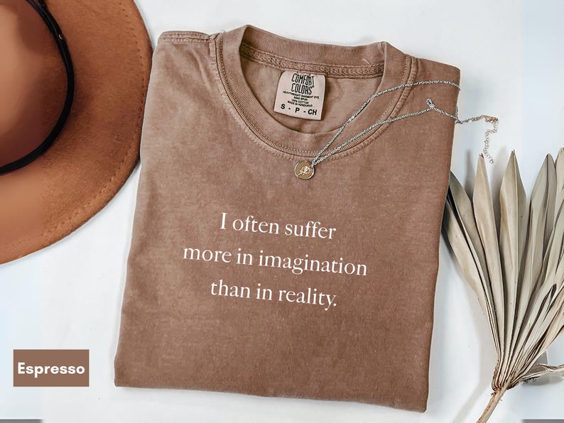 May include: Espresso-colored t-shirt with the quote "I often suffer more in imagination than in reality" in white text. A silver necklace with a pendant and a brown hat are next to the shirt. The shirt has a Comfort Colors tag.