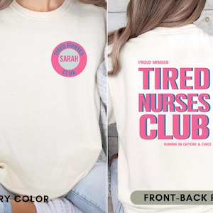 May include: Ivory-colored t-shirt with a front and back print. The front features a pink and blue circular design with the text "Tired Nurses Club" and "Sarah". The back displays "Tired Nurses Club" in pink and blue, with "Running on Caffeine & Chaos".