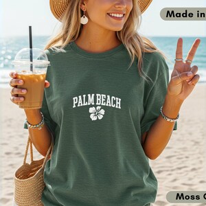 May include: Moss green t-shirt with "PALM BEACH" text and a white flower graphic. The shirt is made in the USA. The person is holding a drink with a straw and a woven tote bag.
