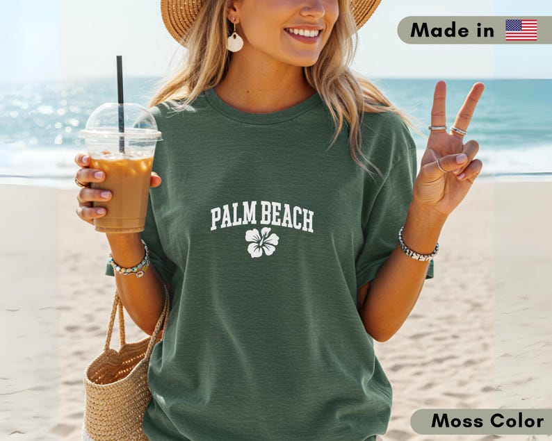 May include: Moss green t-shirt with "PALM BEACH" text and a white flower graphic. The shirt is made in the USA. The person is holding a drink with a straw and a woven tote bag.