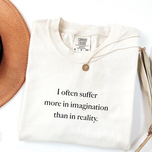 May include: Ivory-colored t-shirt with the text "I often suffer more in imagination than in reality." A gold necklace with a round pendant rests on the shirt. A brown hat and dried plant are in the background.