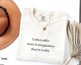 Könnte beinhalten: Elfenbeinfarbenes T-Shirt mit dem Text "I often suffer more in imagination than in reality." Eine goldene Halskette mit rundem Anhänger liegt auf dem Shirt. Ein brauner Hut und eine getrocknete Pflanze sind im Hintergrund.