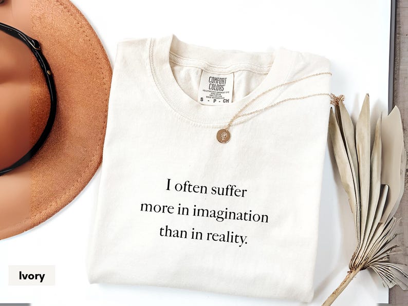 May include: Ivory-colored t-shirt with the text "I often suffer more in imagination than in reality." A gold necklace with a round pendant rests on the shirt. A brown hat and dried plant are in the background.