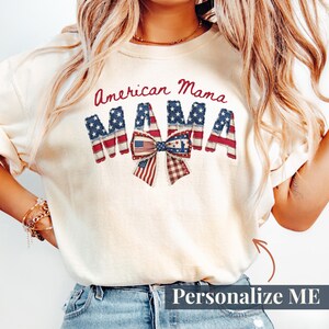 Può includere: T-shirt color crema con la scritta "American Mama" in corsivo rosso. La parola "MAMA" è disegnata con un tema patriottico della bandiera americana, con stelle e strisce. Un fiocco con un disegno della bandiera americana è centrato sotto la parola "MAMA".