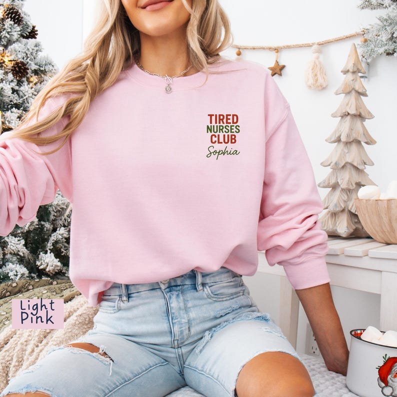 May include: Light pink sweatshirt with the text "Tired Nurses Club Sophia" in red and green. The long-sleeved crew neck top is a solid light pink color. The sweatshirt is a casual style.