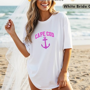 Puede incluir: Camiseta blanca con la frase "CAPE COD" en rosa y un ancla rosa. La camiseta es de corte holgado. Las palabras "White Bride Color" están en un recuadro blanco en la esquina superior derecha. La foto está tomada en la playa.