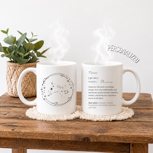 Puede incluir: Dos tazas de cerámica blanca con diseños impresos en negro. Una taza muestra un marco floral circular con la constelación de Piscis y texto. La otra taza muestra una definición de diccionario de Piscis con el nombre Olivia en cursiva.