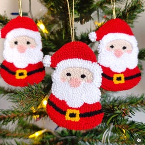 Op de afbeelding: Drie handgemaakte kerstversieringen van vilt in de vorm van de kerstman. Elke versiering is rood en wit met een zwarte riem en een gele gesp. De versieringen hangen aan een groene kerstboom.