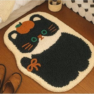 May include: A novelty cat-shaped rug in black and white, featuring an orange apple detail. The rug has green eyes and orange whiskers. It's placed on a wooden floor, with brown leather slippers nearby.