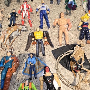 May include: A collection of action figures, including superheroes, a skeleton, and other characters. The figures are in various colors, with some wearing costumes and others appearing muscular. The figures are arranged on a gray surface.