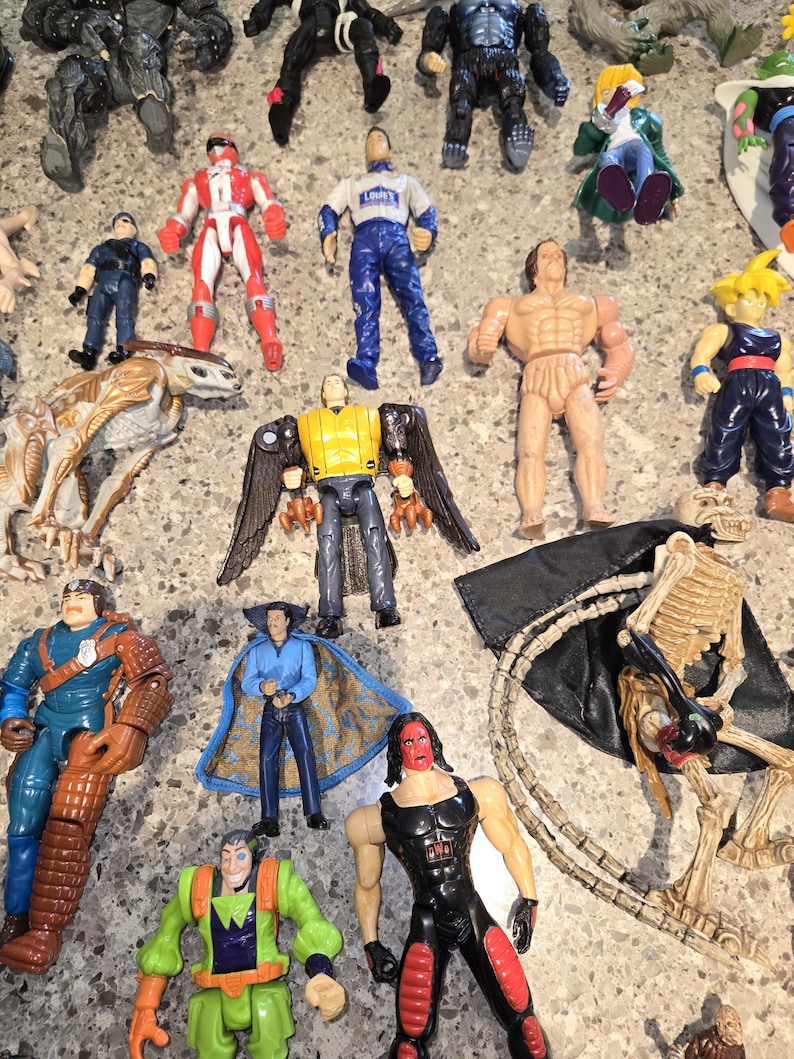 May include: A collection of action figures, including superheroes, a skeleton, and other characters. The figures are in various colors, with some wearing costumes and others appearing muscular. The figures are arranged on a gray surface.