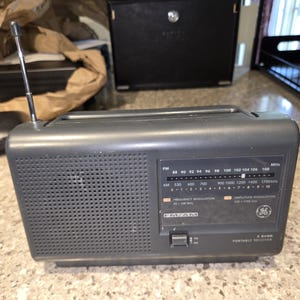 May include: A gray GE portable radio with a telescoping antenna. The radio features an FM/AM dial with frequency markings and a speaker grille. The radio has a rectangular shape and a control panel with a tuning knob and a power switch.
