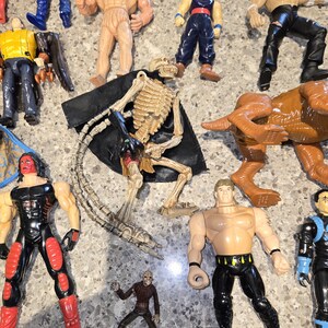 May include: A collection of action figures, including a skeleton figure with a black cape, a brown dinosaur, and several muscular figures in various outfits. The figures are made of plastic and are in different colors.