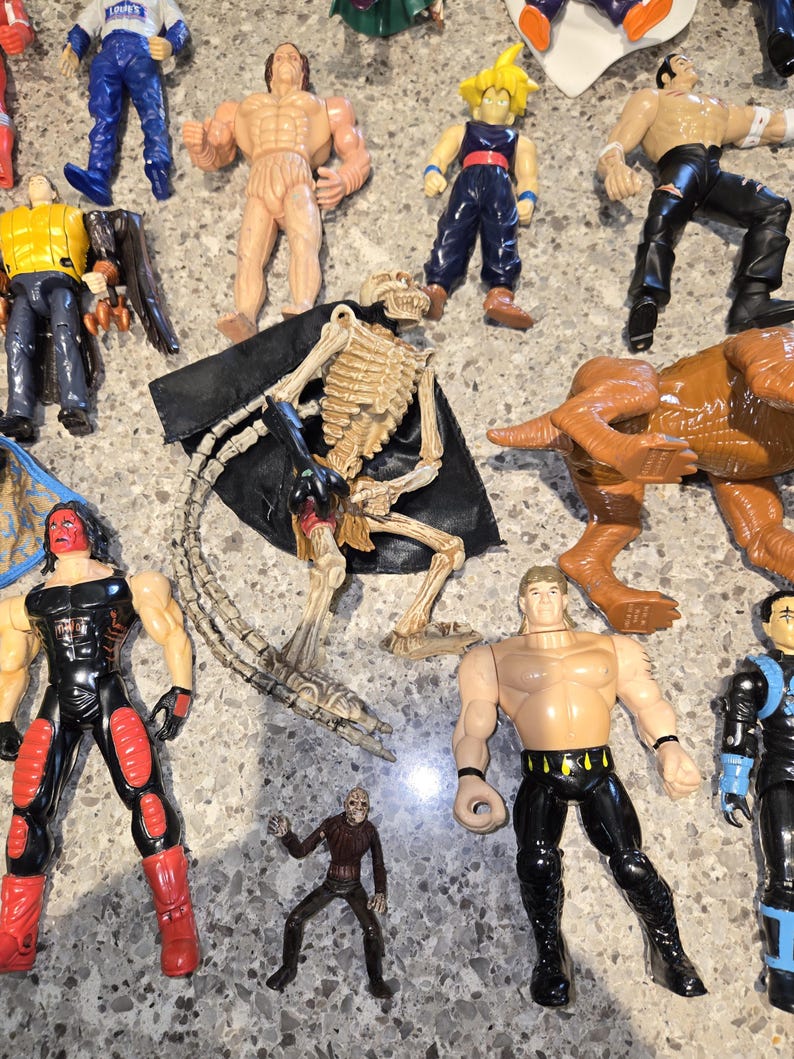 May include: A collection of action figures, including a skeleton figure with a black cape, a brown dinosaur, and several muscular figures in various outfits. The figures are made of plastic and are in different colors.