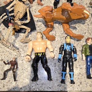 May include: A collection of action figures, including a brown Tyrannosaurus Rex, a skeleton figure with a black cape, and several human-like figures in various outfits. The figures are made of plastic and are in various poses.
