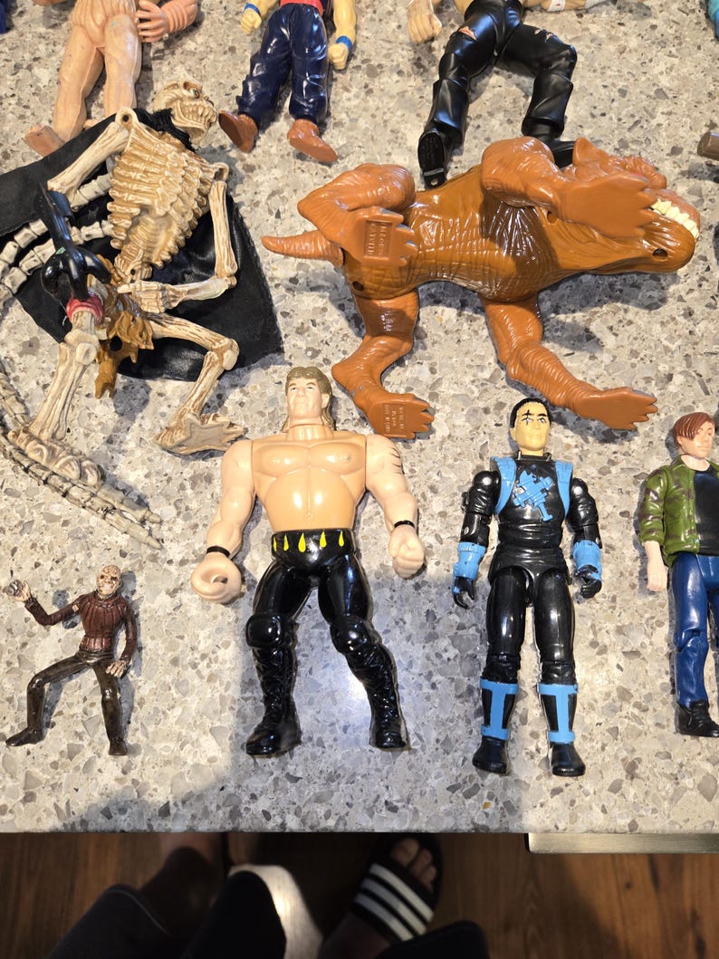 May include: A collection of action figures, including a brown Tyrannosaurus Rex, a skeleton figure with a black cape, and several human-like figures in various outfits. The figures are made of plastic and are in various poses.