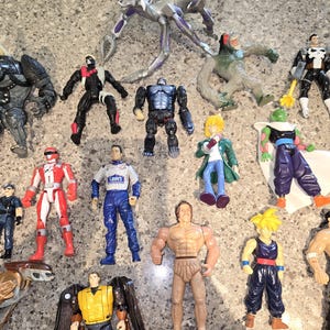 May include: A collection of various action figures, including a silver insect-like creature, a gorilla, a race car driver, and several superheroes. The figures are made of plastic and feature a range of colors and designs.