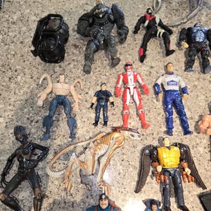 May include: A collection of action figures, including a black armored figure, a red and white Power Ranger, and a blue and white figure with "LOWE'S" on the chest. Other figures include a black alien-like figure and a figure with wings.