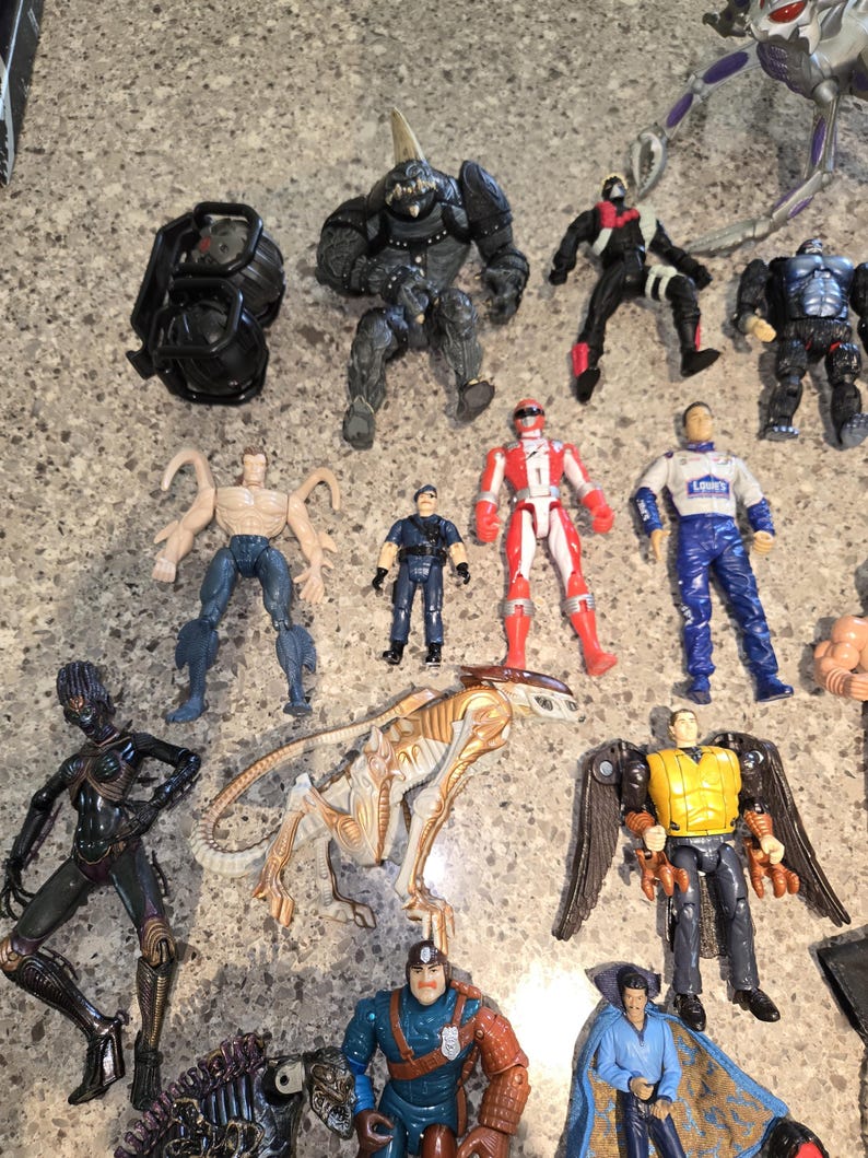 May include: A collection of action figures, including a black armored figure, a red and white Power Ranger, and a blue and white figure with "LOWE'S" on the chest. Other figures include a black alien-like figure and a figure with wings.