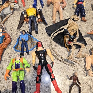 May include: A collection of various action figures, including a skeleton figure with a black cape, a red and black wrestler figure, and a figure with yellow wings. Other figures include a blue-clad figure and a green and purple figure.