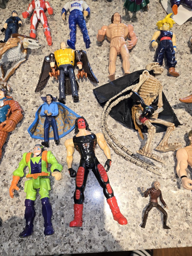 May include: A collection of various action figures, including a skeleton figure with a black cape, a red and black wrestler figure, and a figure with yellow wings. Other figures include a blue-clad figure and a green and purple figure.