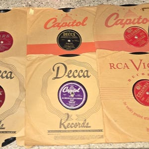 May include: A collection of vintage black vinyl records and their original paper sleeves. The sleeves feature branding from Capitol, Decca, and RCA Victor. The record labels include various colors like red, purple, and black with printed text.