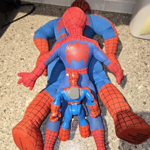 May include: Three Spider-Man toys are arranged on a surface. Two are large, plush figures in red and blue, with the Spider-Man logo on the chest. A smaller, plastic Spider-Man toy is in the center.