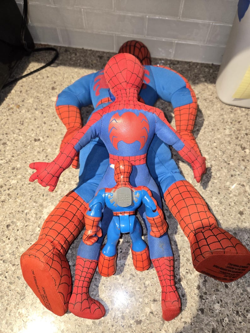 May include: Three Spider-Man toys are arranged on a surface. Two are large, plush figures in red and blue, with the Spider-Man logo on the chest. A smaller, plastic Spider-Man toy is in the center.