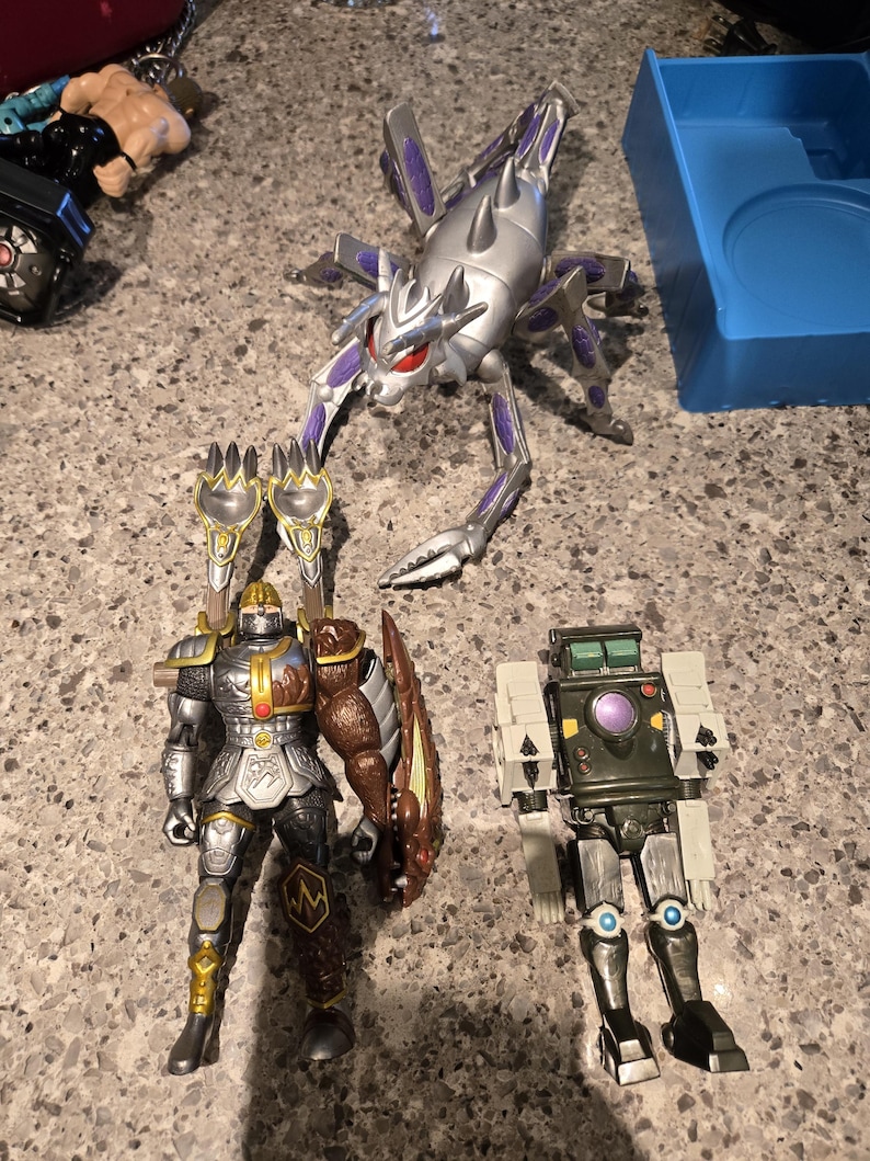 May include: Three action figures including a silver and purple insectoid creature, a knight figure with brown and silver armor featuring two claw-like attachments on its back, and a small dark green and silver robot with a purple circular chest piece.