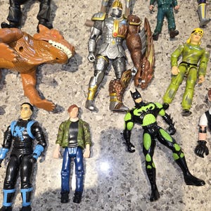 May include: A collection of action figures, including a silver knight, a green and black Batman, and a brown dinosaur. Other figures include characters in blue, green, and yellow outfits. The toys are arranged on a speckled surface.