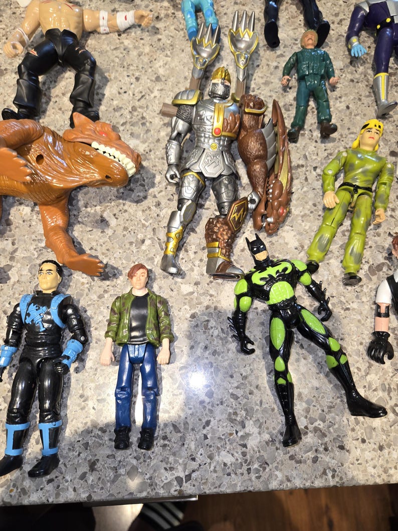 May include: A collection of action figures, including a silver knight, a green and black Batman, and a brown dinosaur. Other figures include characters in blue, green, and yellow outfits. The toys are arranged on a speckled surface.