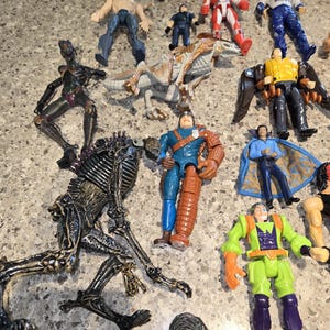 May include: A collection of various action figures, including a dinosaur, a robot, and several human-like figures. The figures are in various colors, including blue, red, green, and black. Some figures have wings or capes.